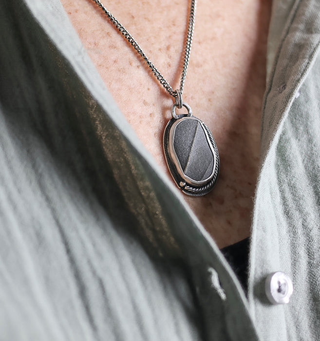 Close-up of a silver pendant necklace with a beach stone on a person wearing a gray shirt.