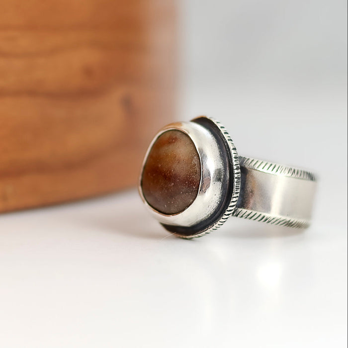 Silver ring with a brown stone on a white surface with a wooden block in the background
