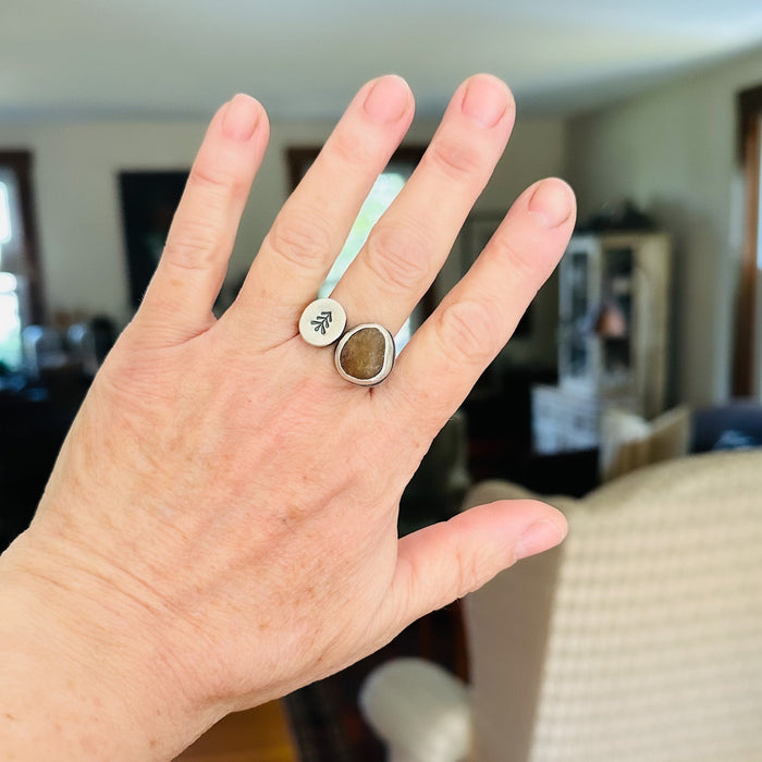 Hand wearing a ring with a brown stone and silver band, set against a blurred indoor background.