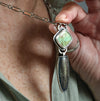 A close-up of a hand holding a silver pendant necklace with a square kingmand turquoise stone with a dangling teardrop-shaped hollow form worn by a person in an olive green shirt.