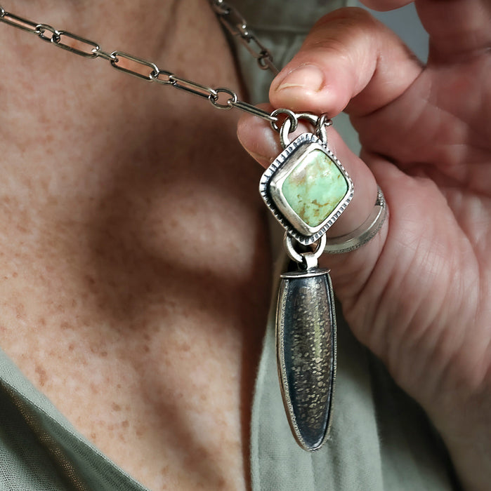 A close-up of a hand holding a silver pendant necklace with a square kingmand turquoise stone with a dangling teardrop-shaped hollow form worn by a person in an olive green shirt.