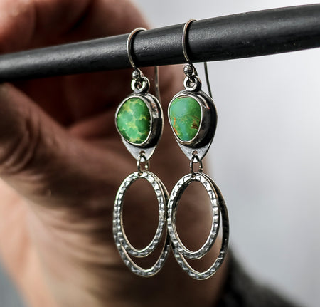 Silver earrings with green turquoise stones hanging from a rod against a blurred background