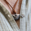 A close-up of a person wearing a  linen shirt and a handmade pendant necklace. The necklace features a brown leather cord, a black beach  stone with a silver plate and grommet