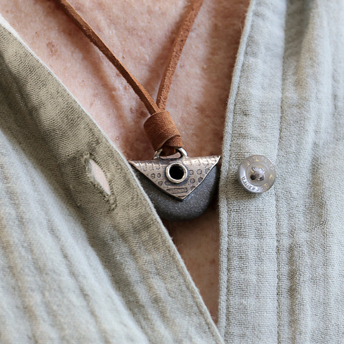A close-up of a person wearing a  linen shirt and a handmade pendant necklace. The necklace features a brown leather cord, a black beach  stone with a silver plate and grommet