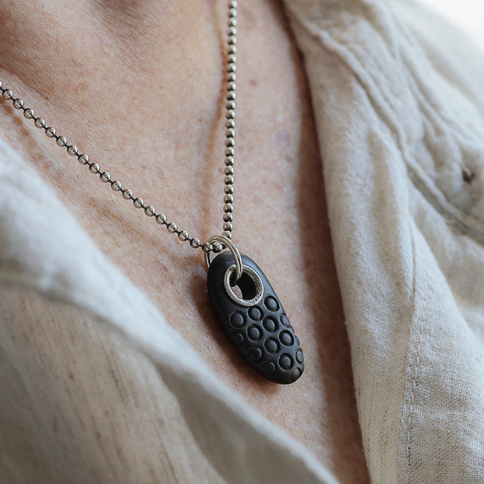 A close-up of a person wearing a silver ball-chain necklace with a dark carved beach stone with a circular design.
