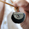 A close-up of a hand holding a black stone pendant necklace with a silver leaf textured plate  and a grommet with tan cord