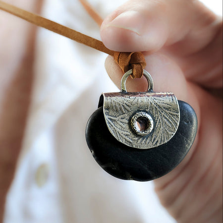 A close-up of a hand holding a black stone pendant necklace with a silver leaf textured plate  and a grommet with tan cord