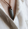 A close-up of a dark gray stone pendant necklace with a brown leather cord, worn over a light beige linen shirt.