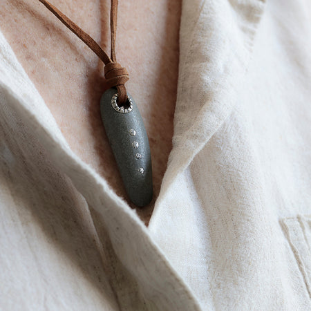 A close-up of a dark gray stone pendant necklace with a brown leather cord, worn over a light beige linen shirt.