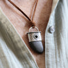 A close-up of a person wearing a pendant necklace with a brown cord, a silver-toned metal cap, and a dark gray stone pendant over a light beige linen shirt.