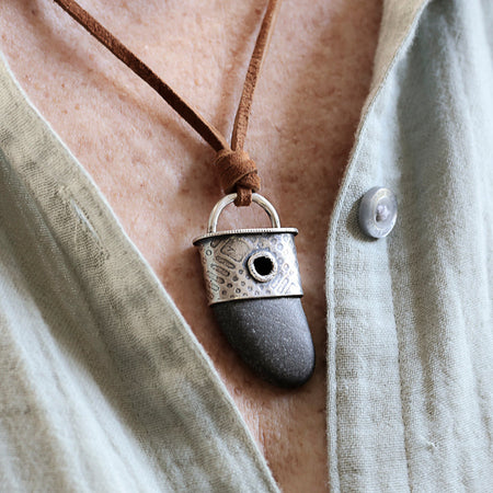 A close-up of a person wearing a pendant necklace with a brown cord, a silver-toned metal cap, and a dark gray stone pendant over a light beige linen shirt.