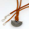 A close-up of a necklace with a gray stone pendant and a silver plate and grommet, suspended on a brown suede cord with silver  hooks at the ends, set against a plain white background.