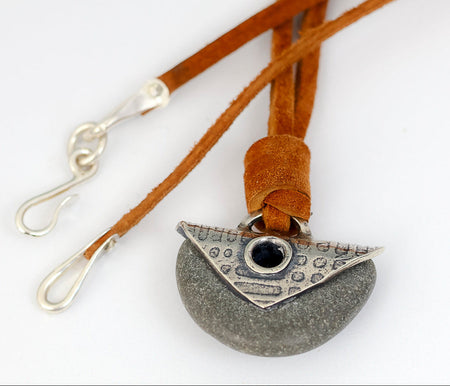 A close-up of a necklace with a gray stone pendant and a silver plate and grommet, suspended on a brown suede cord with silver  hooks at the ends, set against a plain white background.