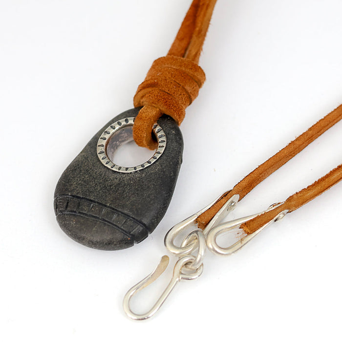 A dark gray stone pendant with a silver grommet threaded onto a tan leather cord with silver clasp, shown on a white background.