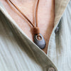 A close-up of a person wearing a dark gray stone pendant necklace with a brown leather cord , styled over a light beige linen shirt.
