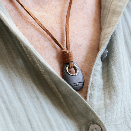A close-up of a person wearing a dark gray stone pendant necklace with a brown leather cord , styled over a light beige linen shirt.