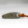 A dark gray stone pendant with three circular cutouts and a brown leather cord knotted through a metal loop, shown on a neutral gray background.