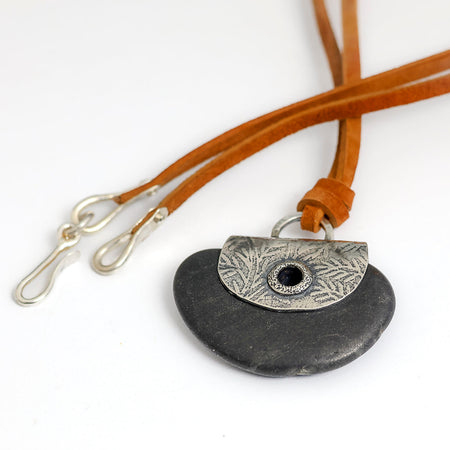 A handmade pendant necklace featuring a dark gray stone disc and a silver textured plate with a grommet, suspended from a brown leather strap with silver clasp hooks.