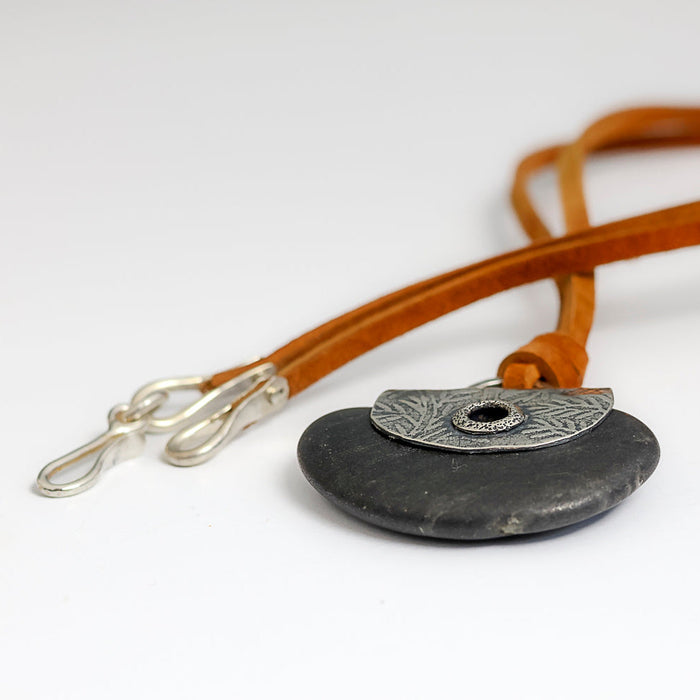 A handmade pendant necklace featuring a dark gray stone disc and a silver textured plate with a grommet, suspended from a brown leather strap with silver clasp hooks.