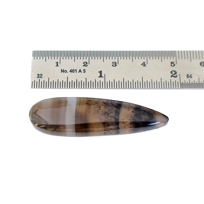 A teardrop-shaped polished agate gemstone with brown and smoky gray banded patterns, shown below a metal ruler for scale on a white background.