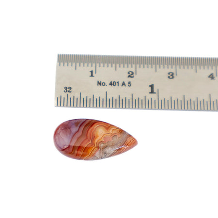 A polished teardrop-shaped  agate cabochon gemstone with orange, red, and white agate-like banding, shown next to a metal ruler for scale on a white background.