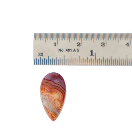 A teardrop-shaped  agate cabochon gemstone with concentric bands of orange, red, brown, and cream, shown below a metal ruler for scale on a white background.