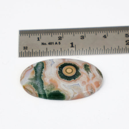 An oval jasper cabochon gemstone with a natural  pattern in green, peach, cream, and gold, shown below a metal ruler for scale on a white background.