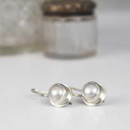 A pair of sterling silver earrings with round iridescent coin pearls, displayed on a white surface with a softly blurred glass jar in the background.