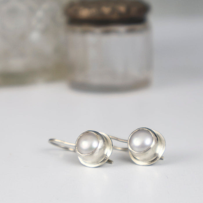 A pair of sterling silver earrings with round iridescent coin pearls, displayed on a white surface with a softly blurred glass jar in the background.