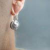 A close-up side view of a person's ear wearing a silver dangle earring with a round  white pearl.