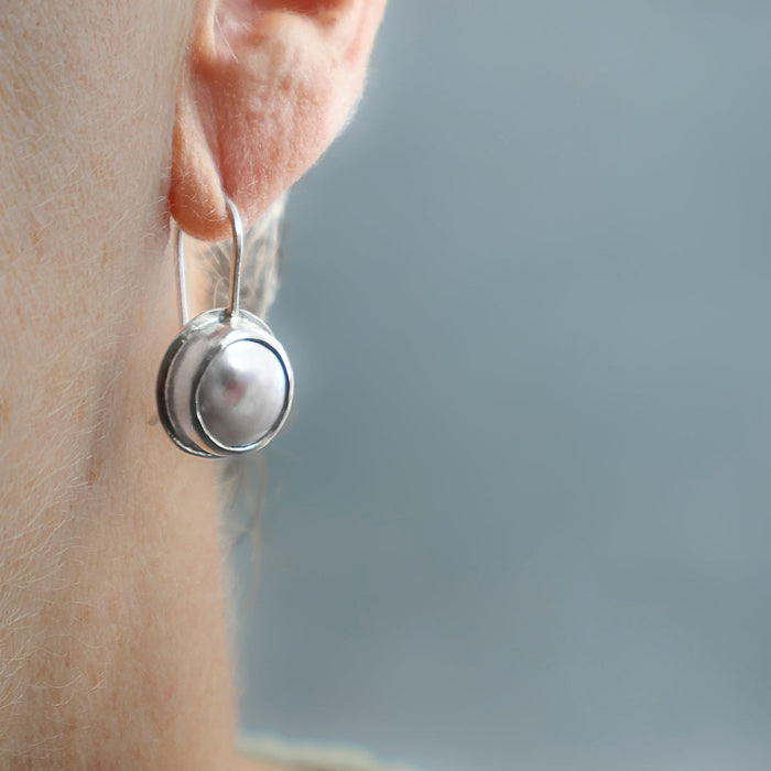 A close-up side view of a person's ear wearing a silver dangle earring with a round  white pearl.