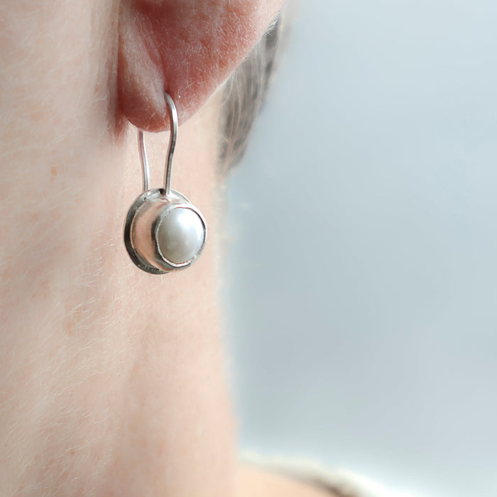 A close-up side view of a person's ear wearing a silver dangle earring with a round  white pearl.