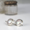 A pair of sterling silver earrings with round iridescent coin pearls, displayed on a white surface with a softly blurred glass jar in the background.