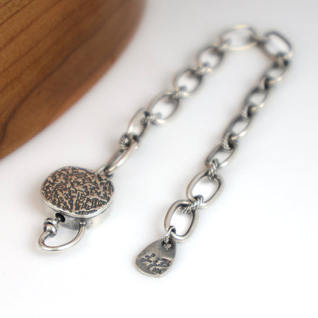A silver chain bracelet with a textured round clasp, photographed on a white surface with a wooden curved prop at the upper left.