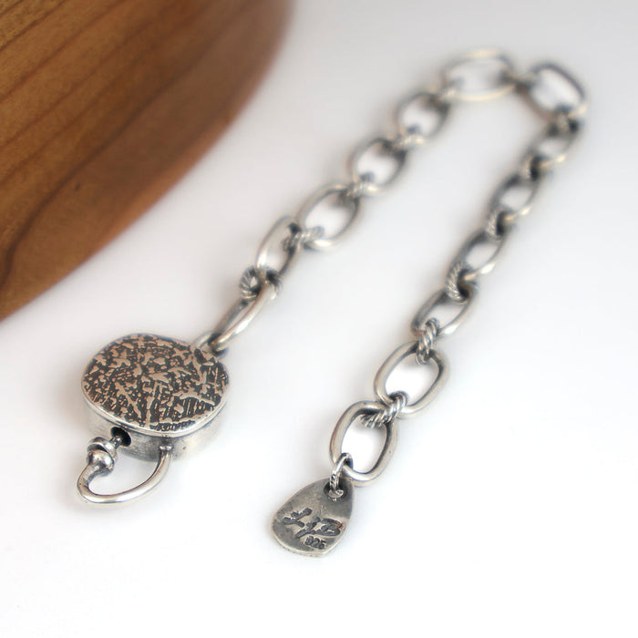 A silver chain bracelet with a textured round clasp, photographed on a white surface with a wooden curved prop at the upper left.