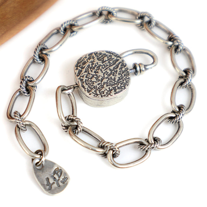 A silver chain bracelet with elongated oval links, twisted rope details at the link joints, a round toggle,  displayed on a white background with a wooden surface in the corner.