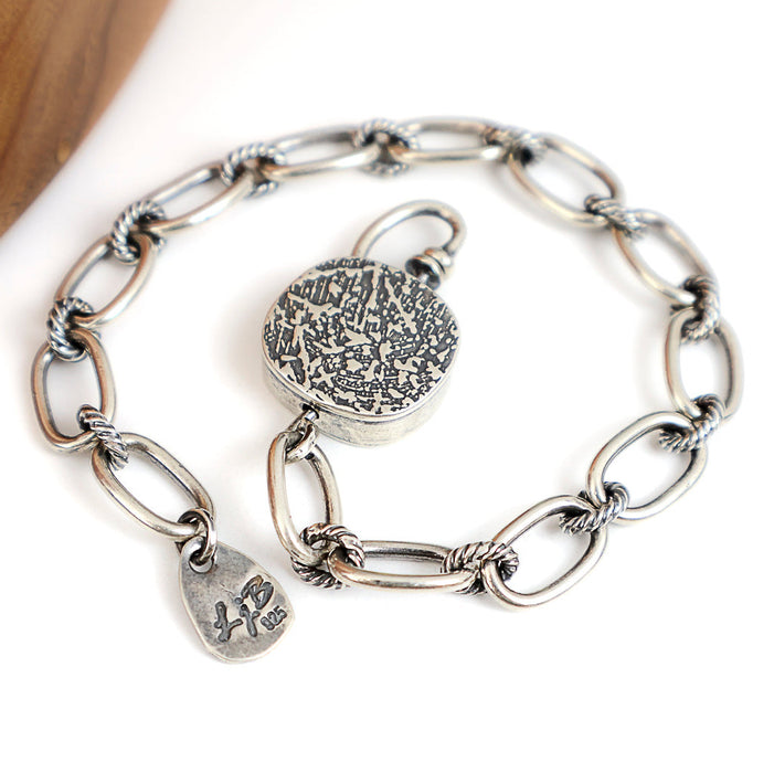 A silver chain bracelet with textured oval links, a round textured toggle, displayed on a clean white background.