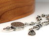 A close-up of a silver chain bracelet with a round textured toggle, resting on a white surface with a blurred wooden object in the background.