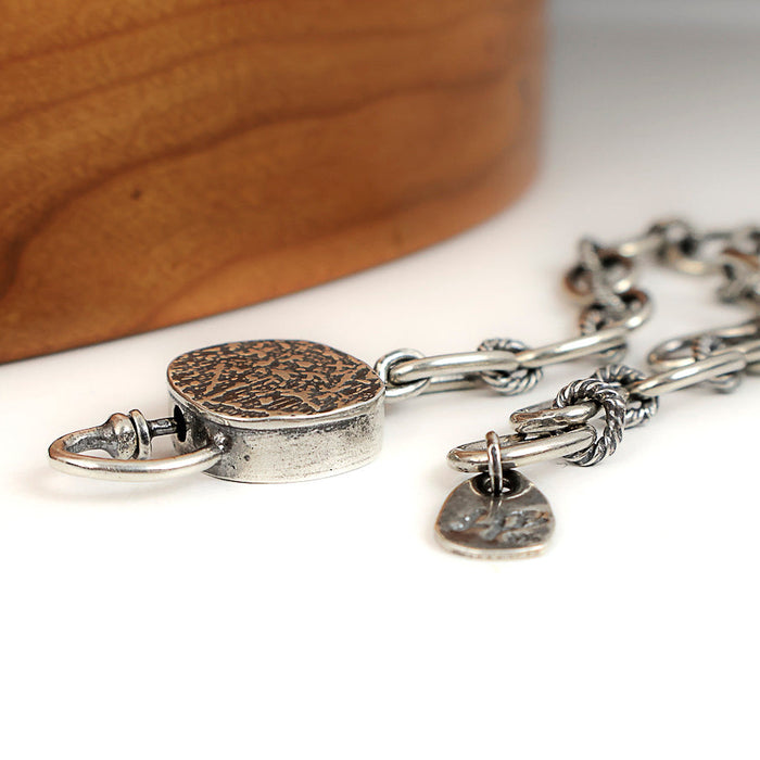 A close-up of a silver chain bracelet with a round textured toggle, resting on a white surface with a blurred wooden object in the background.