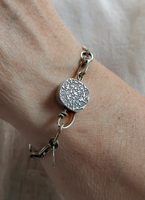 A close-up of a silver bracelet worn on a wrist, featuring a textured toggle and delicate chain links, set against a soft beige fabric background.