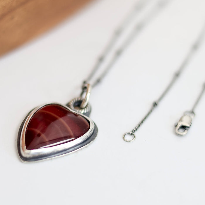 Heart-shaped pendant with red stone on a silver chain against a white background
