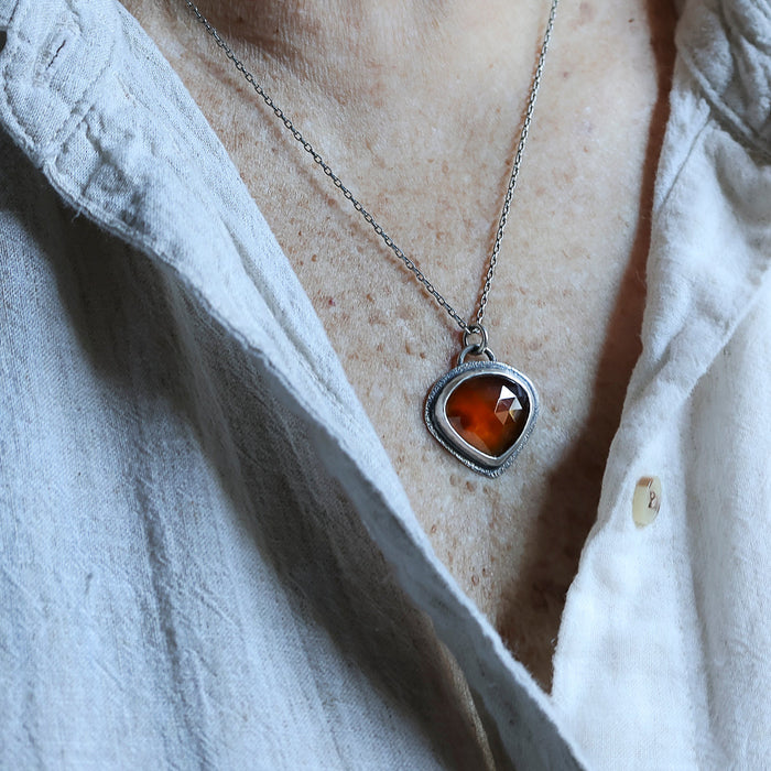 Handmade sterling silver pendant  worn on a person with dark orange rose cut hessonite garnet set point down, sand textured border, on an oval rolo chain