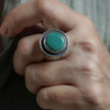 A close-up of a hand wearing a large silver ring with a round turquoise-colored stone.