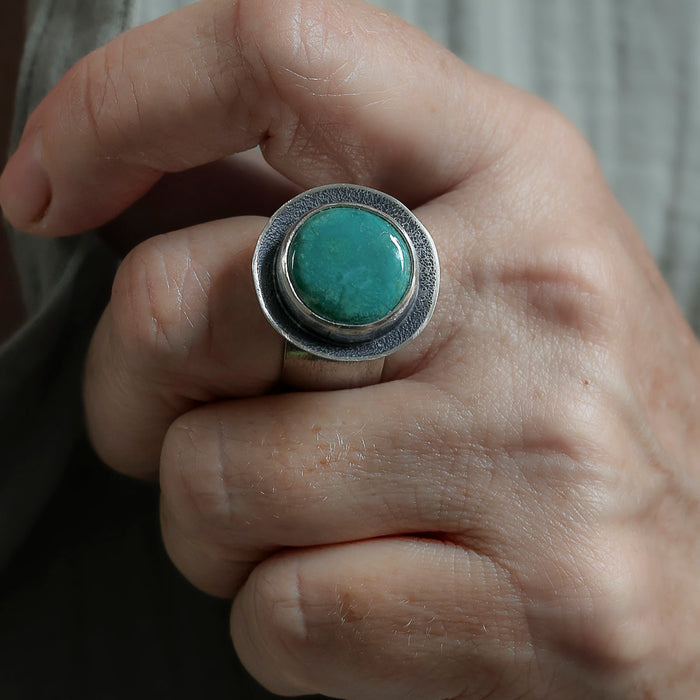A close-up of a hand wearing a large silver ring with a round turquoise-colored stone.