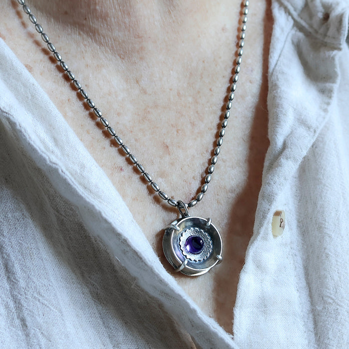 Handmade sterling silver pendant worn by a person with high dome amethyst in a sand textured flower frame inside a pronged silver cup, on an oval bead chain