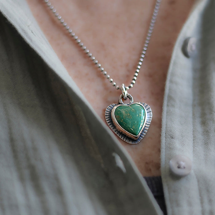 Necklace with a heart-shaped green turquoise pendant worn by a person in a light-colored shirt.