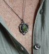 A close-up of a sterling silver necklace with a turquoise stone, worn on a person's neck over an olive green button-up shirt.