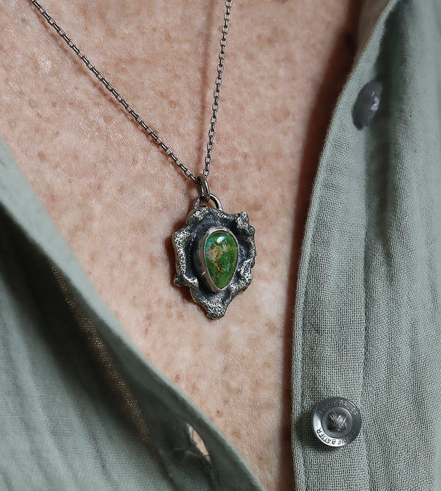 A close-up of a sterling silver necklace with a turquoise stone, worn on a person's neck over an olive green button-up shirt.