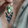 A close-up of a hand holding a silver pendant necklace with a green turquoise stone and textured metal details, against a blurred background of skin and a light-colored shirt.