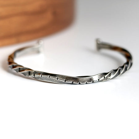 Silver twisted cuff bracelet on a white surface with a wooden object in the background